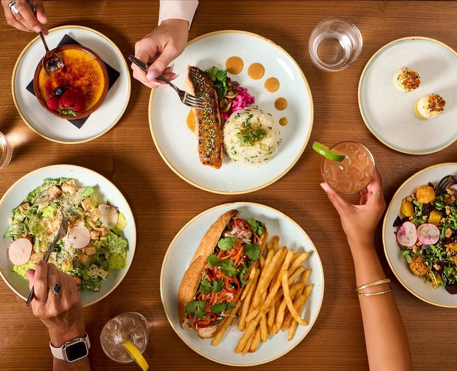 Dishes of food on a table with hands grabbing for drinks and forks