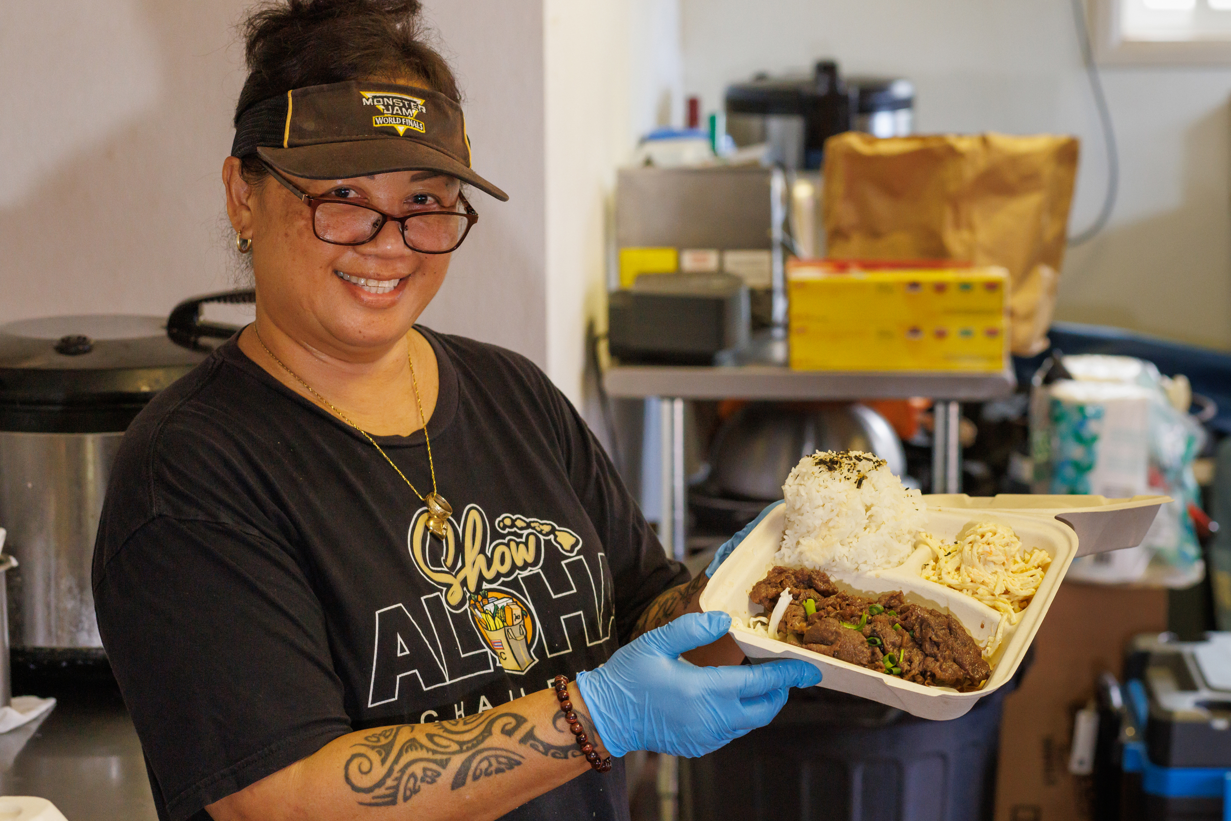 A woman in a visor and glasses holding a plate lunch. Hawaiian foods.